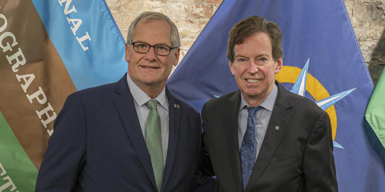 Two men in suits pose in front of flags