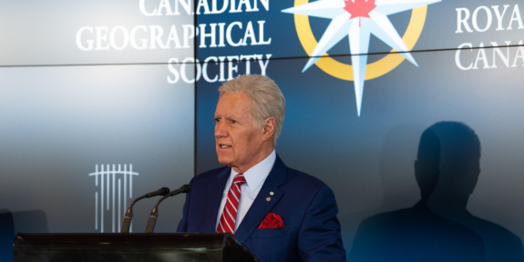 Alex Trebek stands at a podium behind the Royal Canadian Geographical Society logo