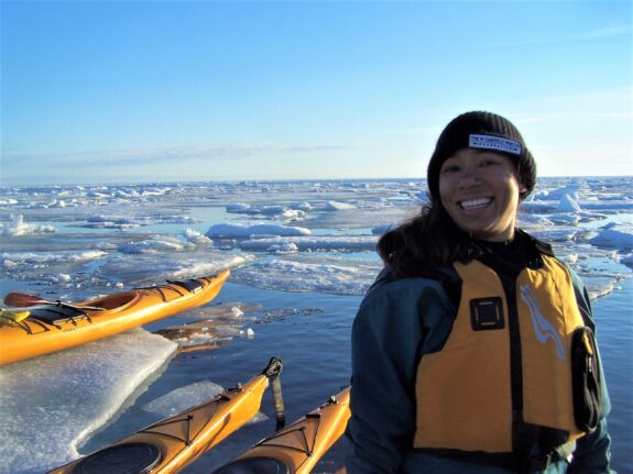 Emily Choy stands in front of an Arctic landscape in a yellow lifejacket alongside yellow kayaks